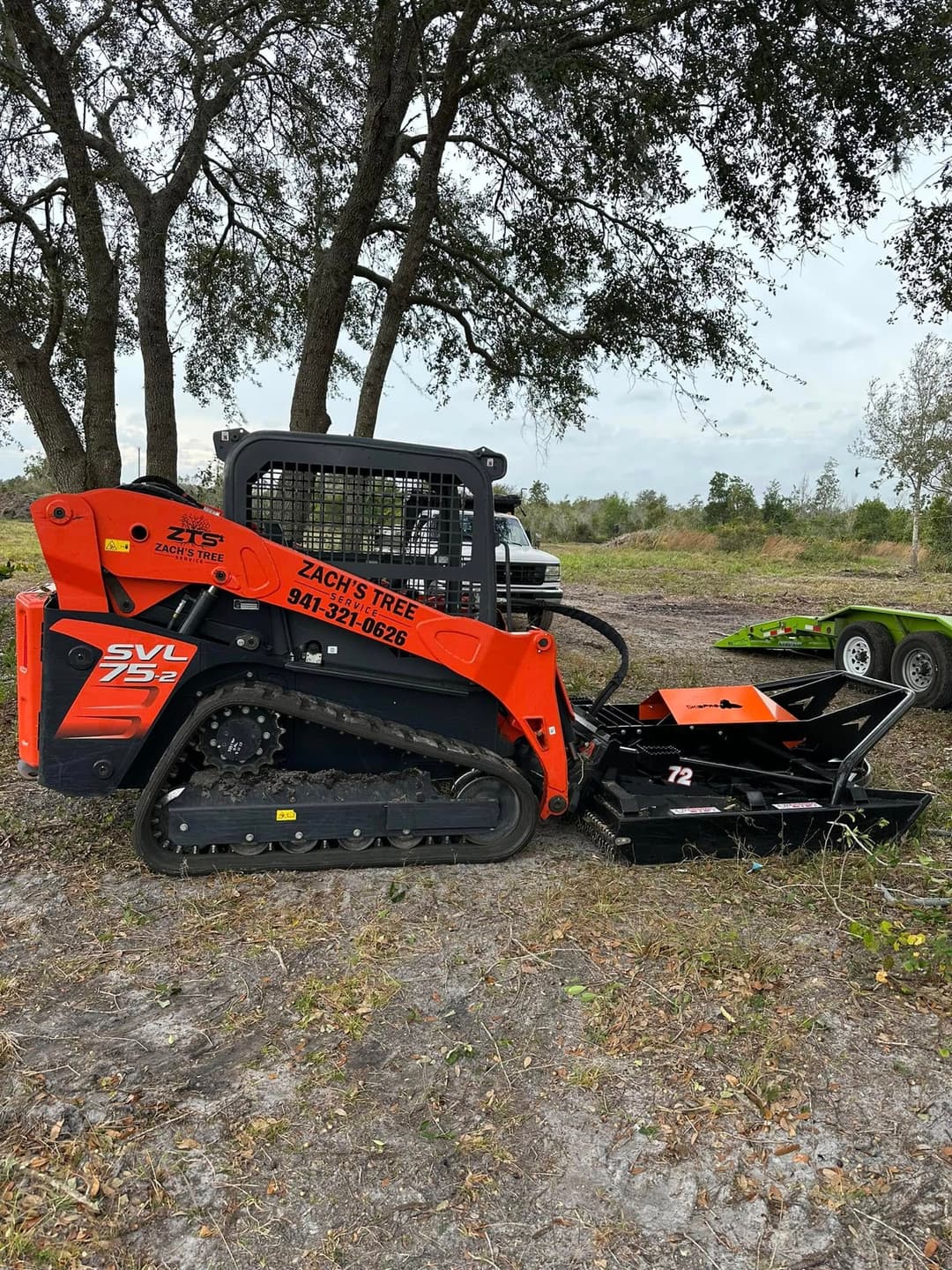 Zach's Tree Service compact track loader with brush cutter attachment in a wooded area.